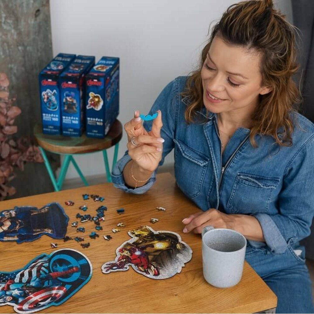Donna tiene un pezzo di puzzle. Camicia di jeans. Pezzi di puzzle su un tavolo. Tazza grigia. Puzzle Captain America e Iron Man. Scatole di puzzle sullo sfondo.