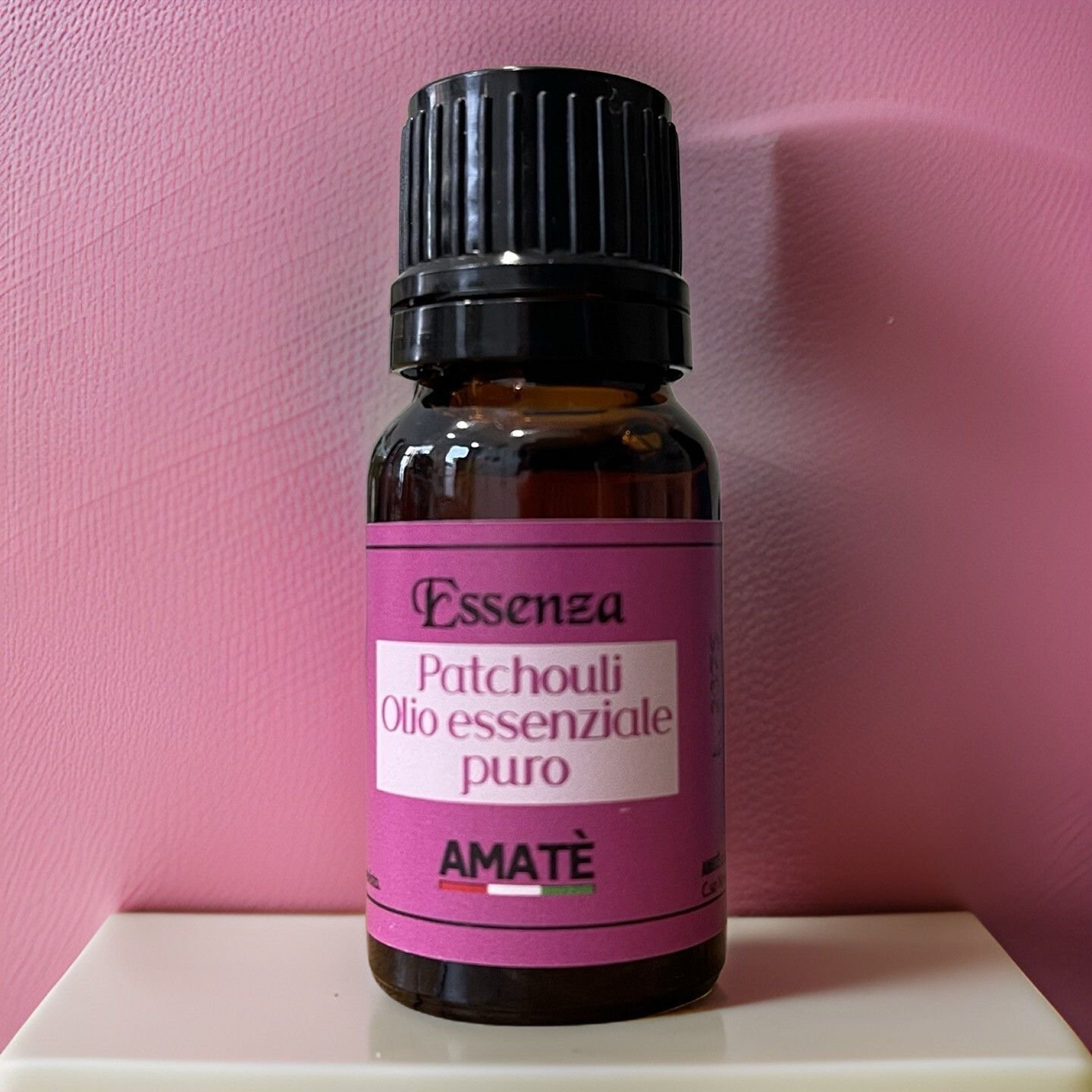 Flacone di vetro marrone con tappo nero. Etichetta con testo: Essenza Patchouli Olio essenziale puro. Marchio: AMATÈ. Su sfondo rosa.