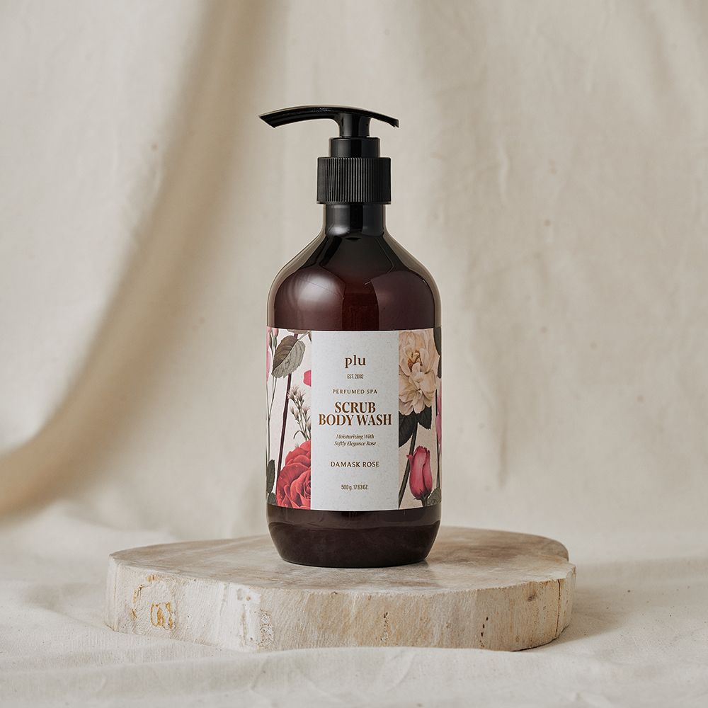 Flacone marrone con dosatore nero. Etichetta floreale con testo "SCRUB BODY WASH DAMASK ROSE". Su un piedistallo.