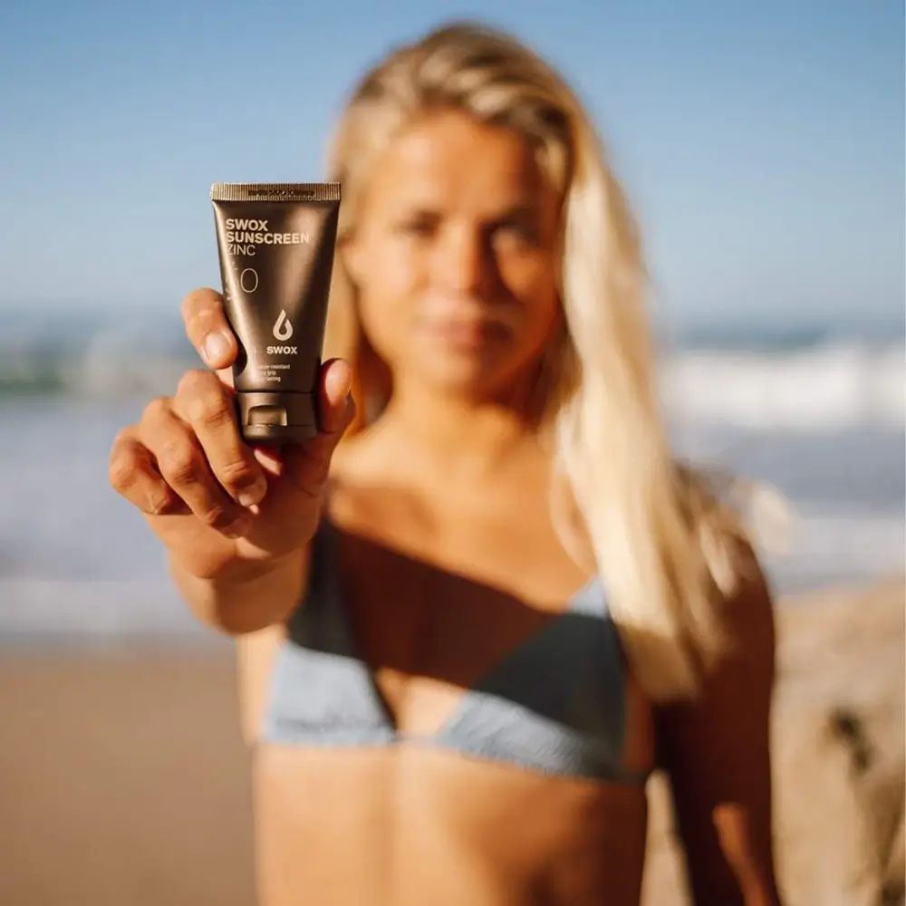 Persona tiene in mano un tubetto di crema solare. Scritta: SWOX SUNSCREEN ZINC. Tubetto marrone, testo nero.