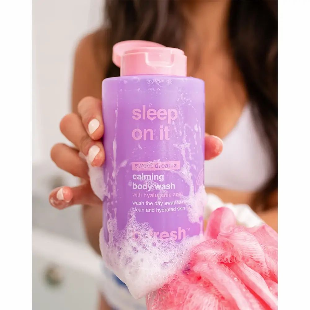 Flacone di gel doccia viola con tappo rosa. Testo: "sleep on it", "calming body wash". Il prodotto è tenuto da una persona, schiuma intorno.