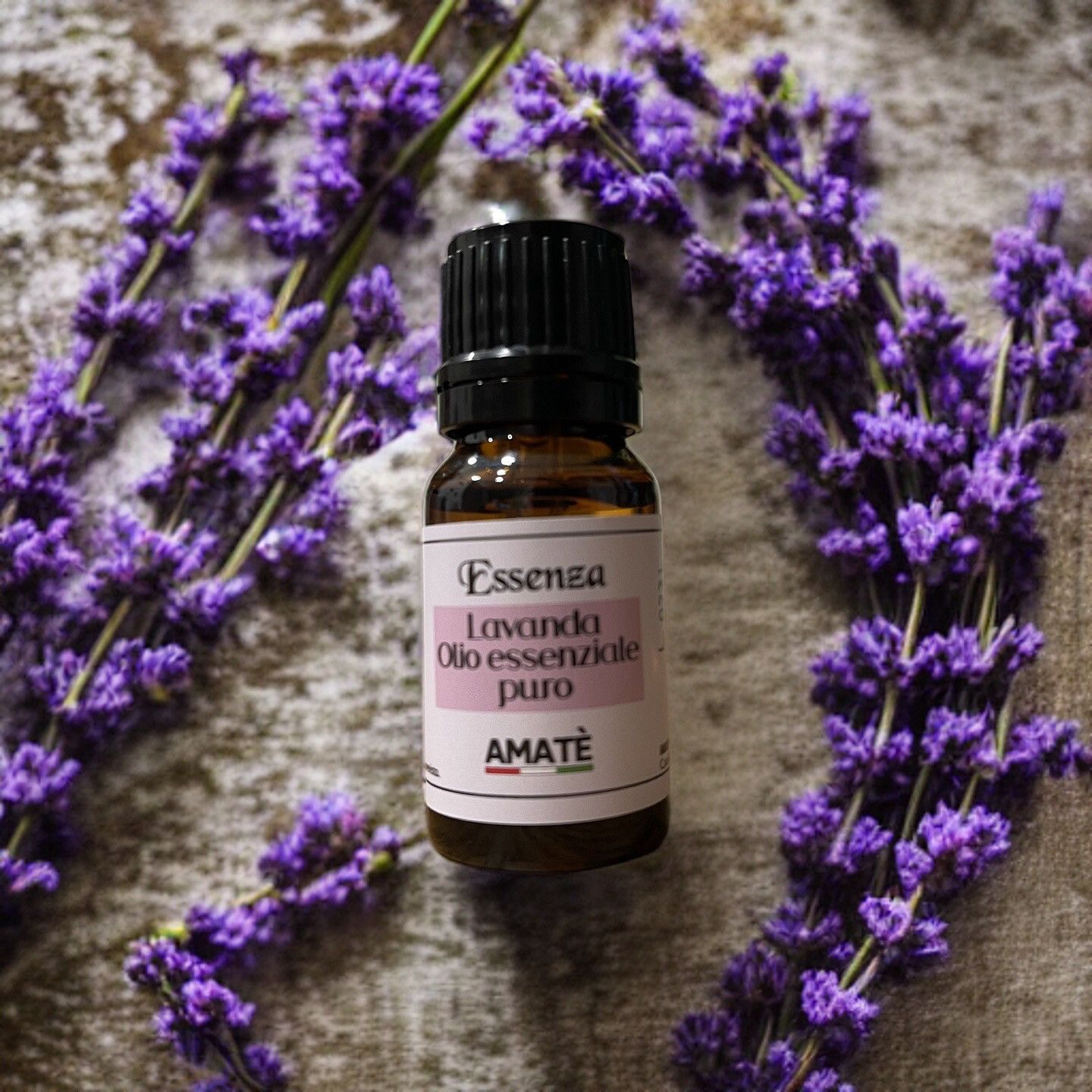 Flacone di vetro marrone con tappo nero, circondato da fiori di lavanda. Etichetta con testo: Essenza Lavanda Olio essenziale puro. Marchio: AMATÈ.