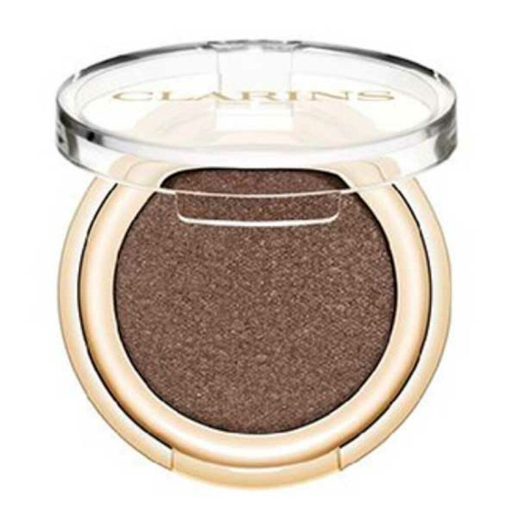 Contenitore rotondo dorato per ombretto con coperchio trasparente. L'ombretto è marrone. Il marchio Clarins è visibile sul coperchio.