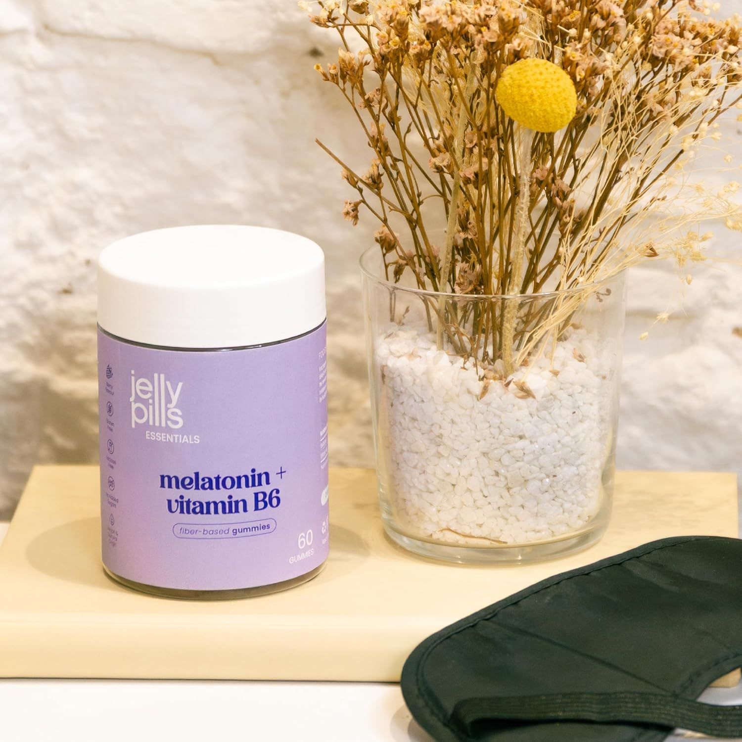 Jelly Pills Essentials Melatonina + Vitamina B6. Barattolo viola con 60 caramelle gommose. Su una tavola di legno. Accanto a un vaso e una mascherina per dormire.