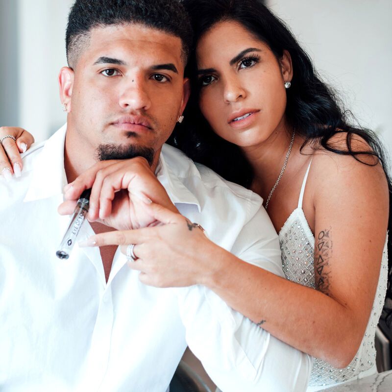 Un uomo e una donna guardano la fotocamera. L'uomo tiene in mano un flacone di profumo. Scritta: EYE OF LOVE Pheromone Parfum Romantic.