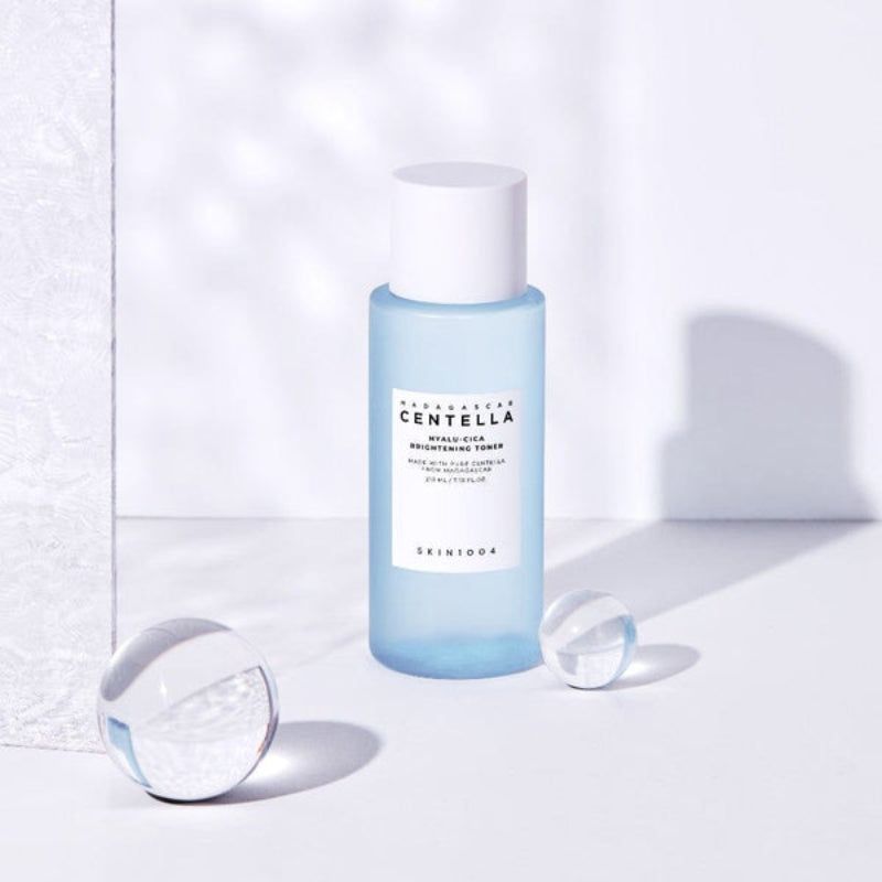 Flacone blu chiaro con tappo bianco. Testo: CENTELLA, HYALU-CICA, Brightening Toner. SKIN1004. Due sfere.