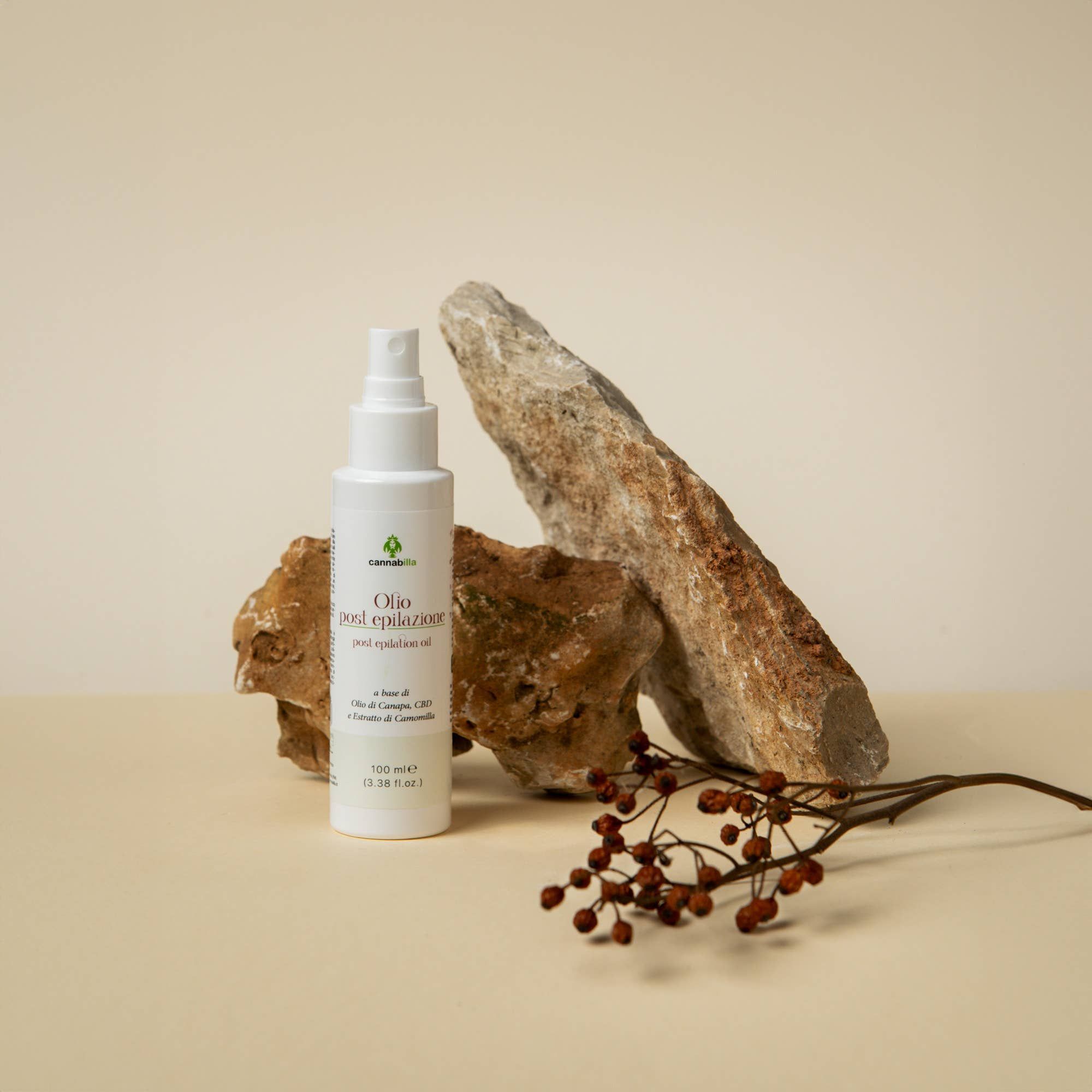 Flacone spray bianco con etichetta su pietre. Scritta: Olio post epilazione. Accanto: ramo con bacche rosse. Sfondo: beige.