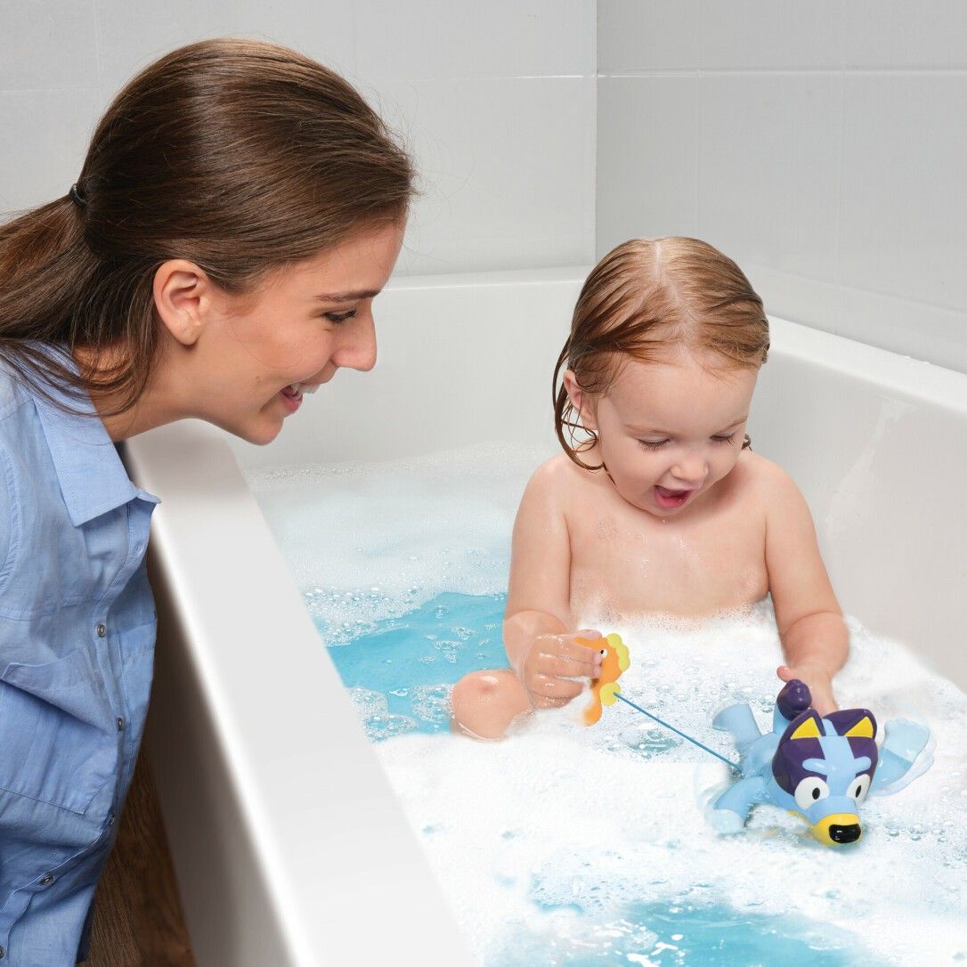 Bambino e donna giocano con il giocattolo da bagno Bluey in una vasca. Acqua e schiuma visibili.