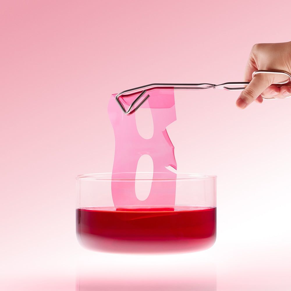 Maschera idrogel rosa sollevata da un liquido rosso con pinzette.