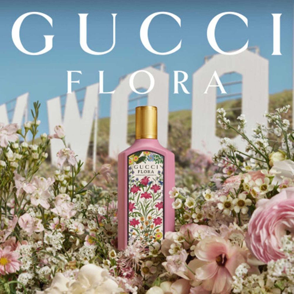 Flacone rosa Gucci Flora con tappo dorato, circondato da fiori. Sullo sfondo: insegna di Hollywood. Testo: Gucci Flora.