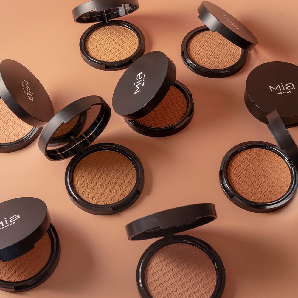 Diversi contenitori cosmetici rotondi aperti con polvere marrone. Coperchi neri con logo Mia Makeup. Su sfondo marrone.