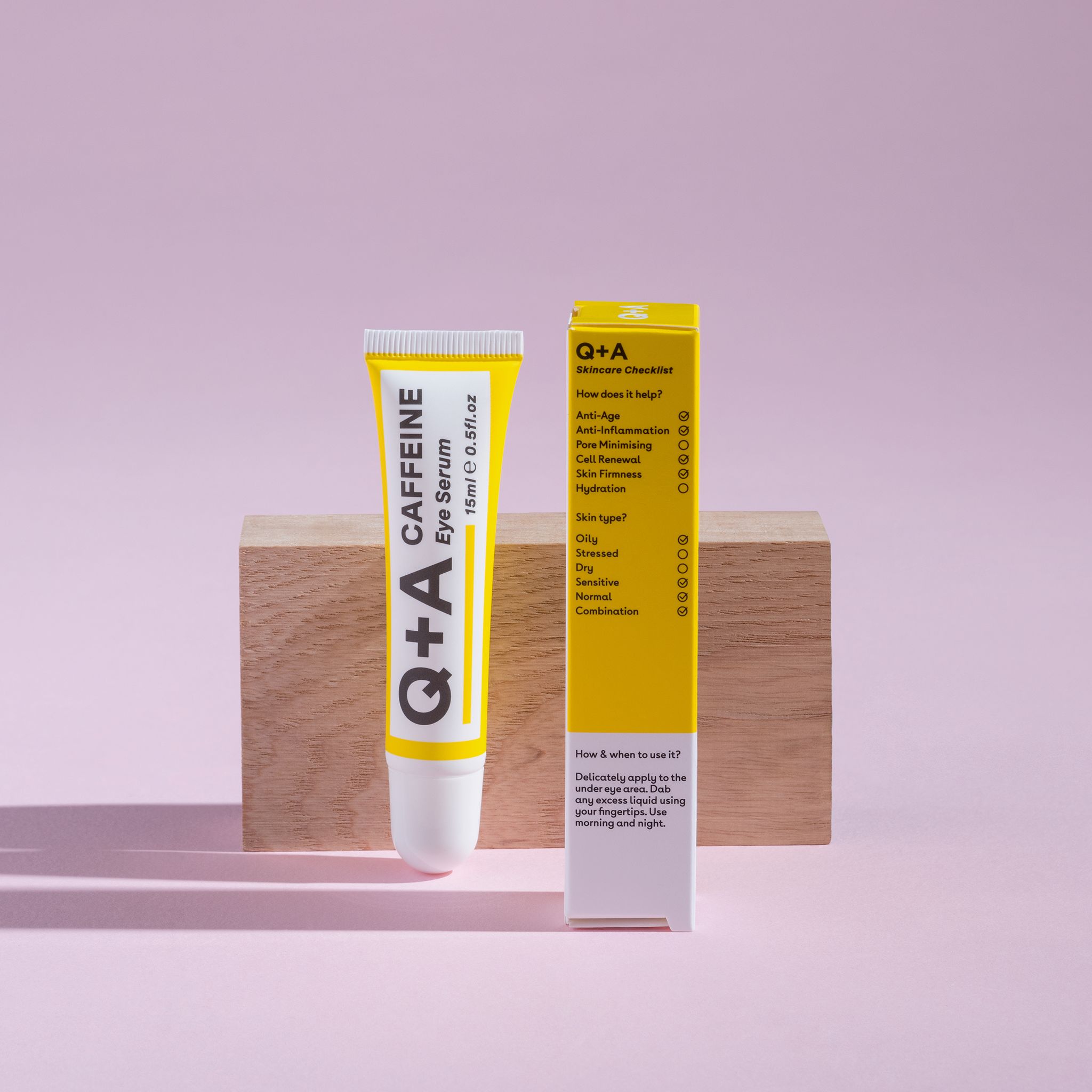 Tubo e scatola gialli. Scritta: Q+A, CAFFEINE Eye Serum, 15ml 0.5fl.oz. Su sfondo rosa con blocco di legno.