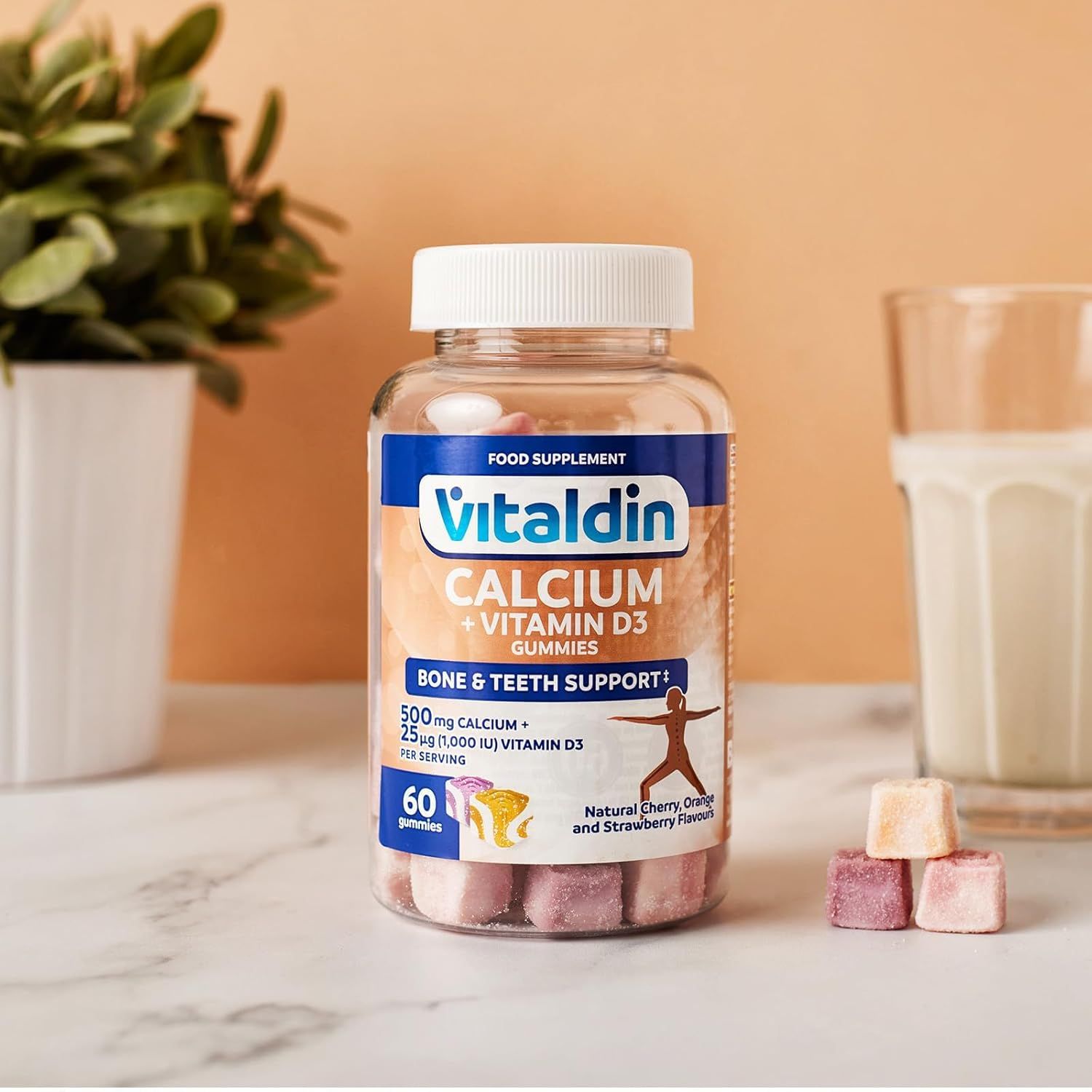 Flacone VITALDIN Calcium + Vitamina D3 Gummies. 60 caramelle gommose. Contiene 500mg calcio e 25µg vitamina D3. Caramelle gommose accanto. Bicchiere di latte.