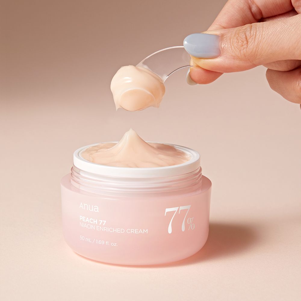 Una mano preleva la crema arricchita alla niacinamide ANUA Peach 77% con una spatola da un vasetto rosa.