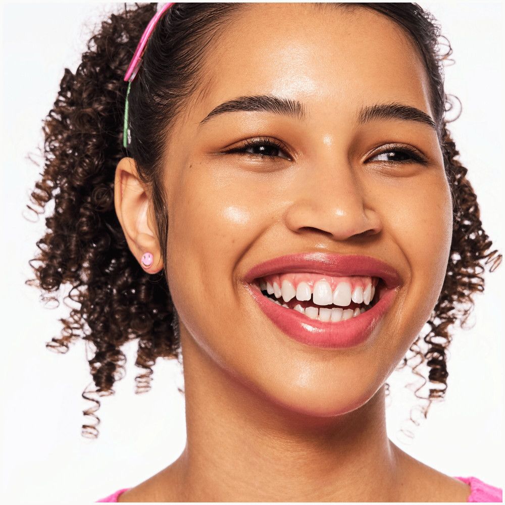 Primo piano di un viso sorridente. Rossetto rosa. Capelli ricci. Orecchino rosa. Sfondo chiaro.