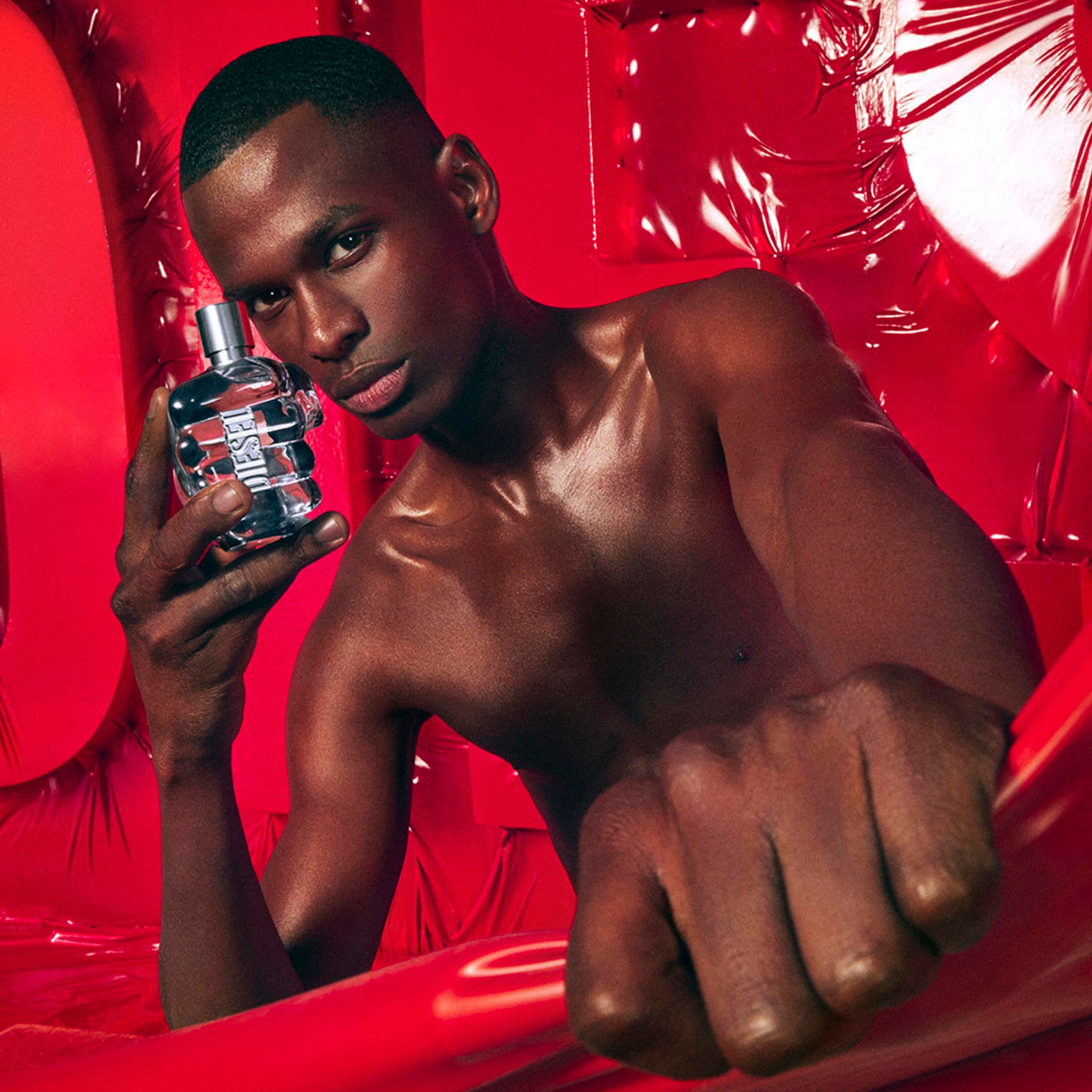 Uomo tiene flacone Diesel Only The Brave. Flacone a forma di pugno, vetro blu. Sfondo rosso.