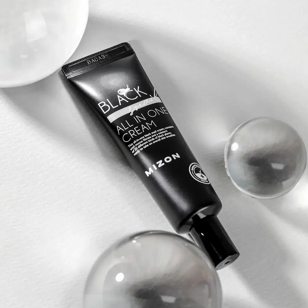 Tubo nero con scritta BLACK Snail ALL IN ONE CREAM e MIZON. Su sfondo bianco con sfere di vetro.
