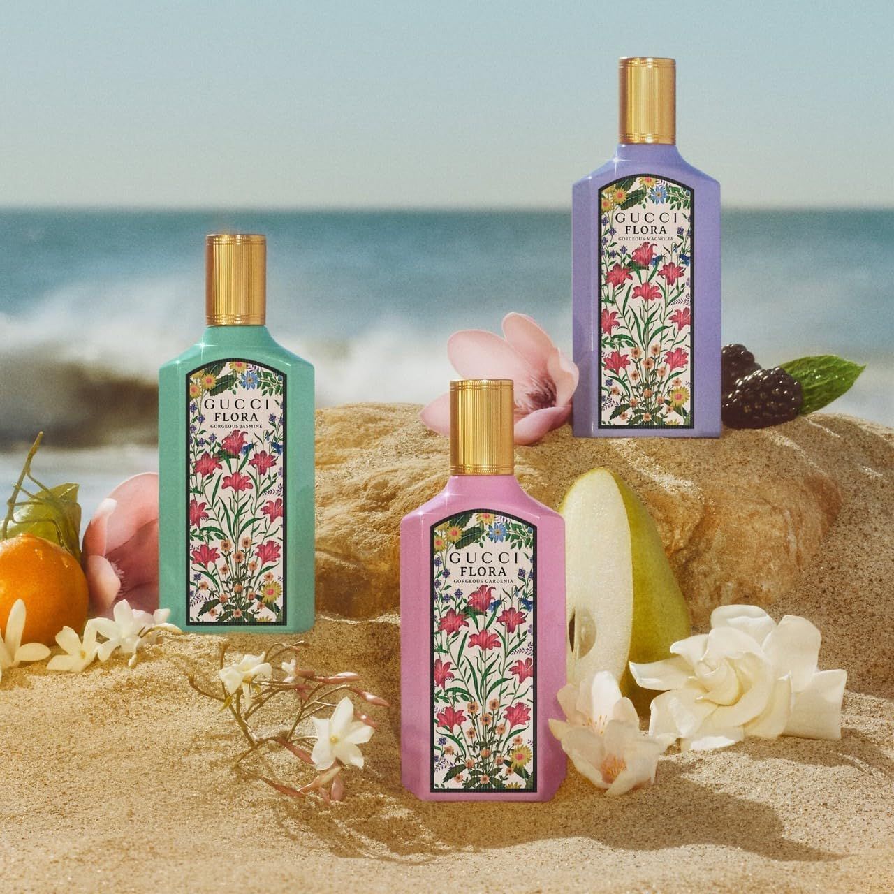 Tre flaconi con etichette floreali. Tappi dorati. Scritta: Gucci Flora Gorgeous Magnolia. Sfondo spiaggia.