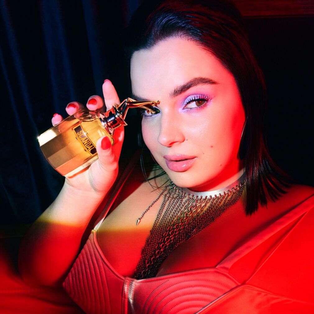 Donna tiene flacone di profumo con tappo dorato. Flacone con scritta Scandal. La donna indossa top rosso e gioielli.