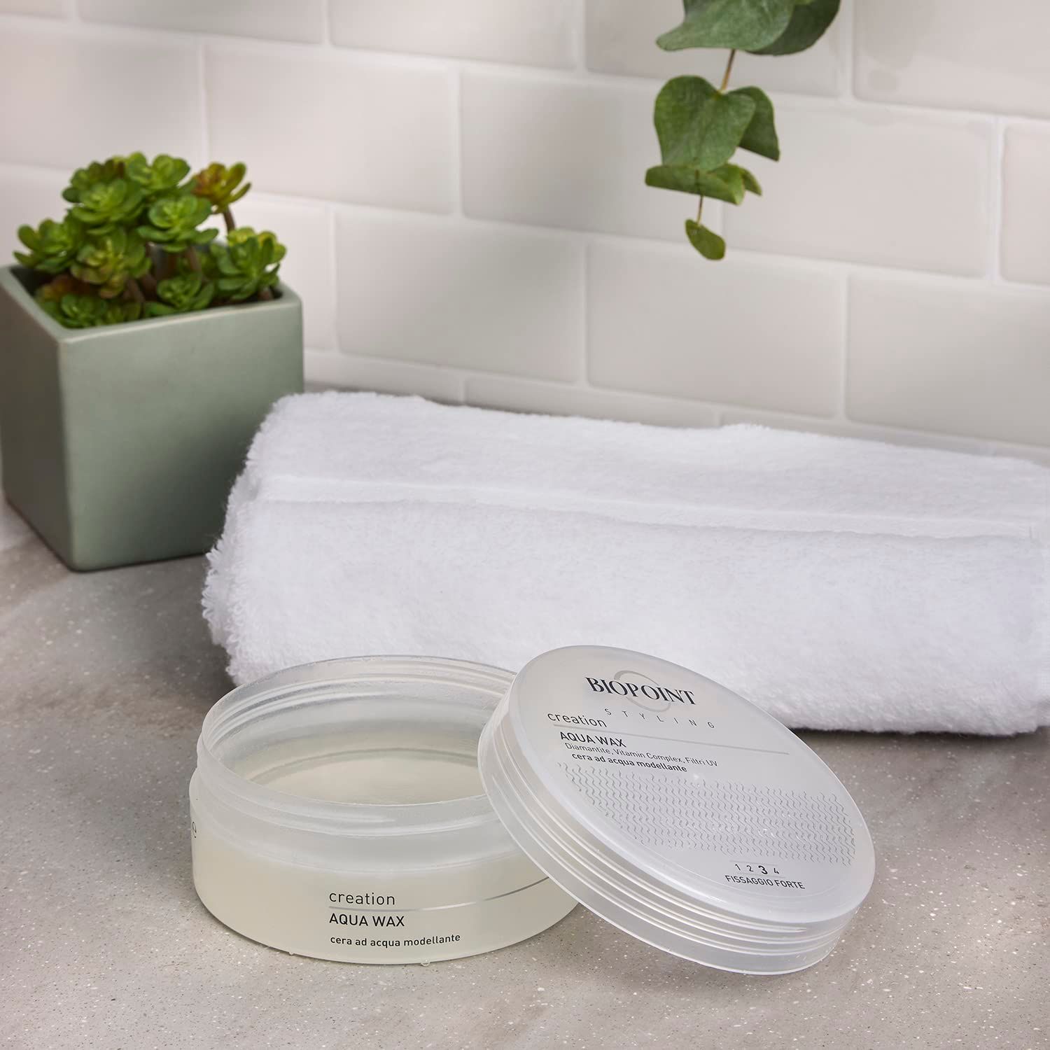 Barattolo trasparente aperto e coperchio. Scritta "BIOPOINT creation AQUA WAX". Su una superficie, accanto ad un asciugamano e una pianta.