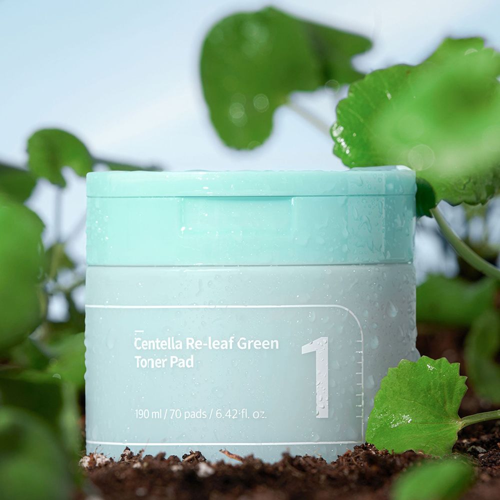 Barattolo verde chiaro con coperchio, circondato da terra e foglie. Scritta: Centella Re-leaf Green Toner Pad, 1. Gocce d'acqua.