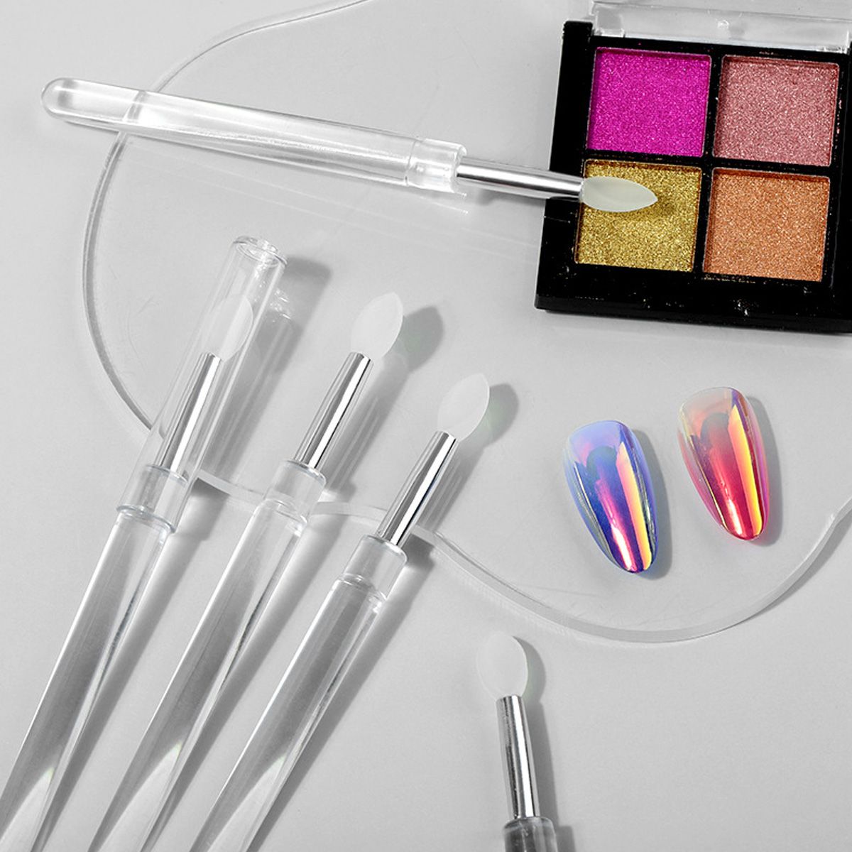 Diversi applicatori trasparenti con punte in silicone accanto a un quad di ombretti e unghie olografiche.