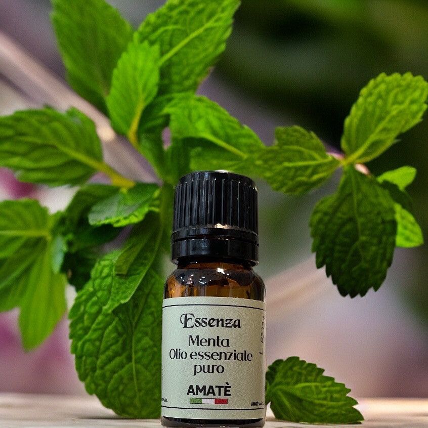 Flacone di vetro marrone con tappo nero, circondato da foglie di menta. Etichetta: Essenza Menta Olio essenziale puro. Marchio: AMATÈ.