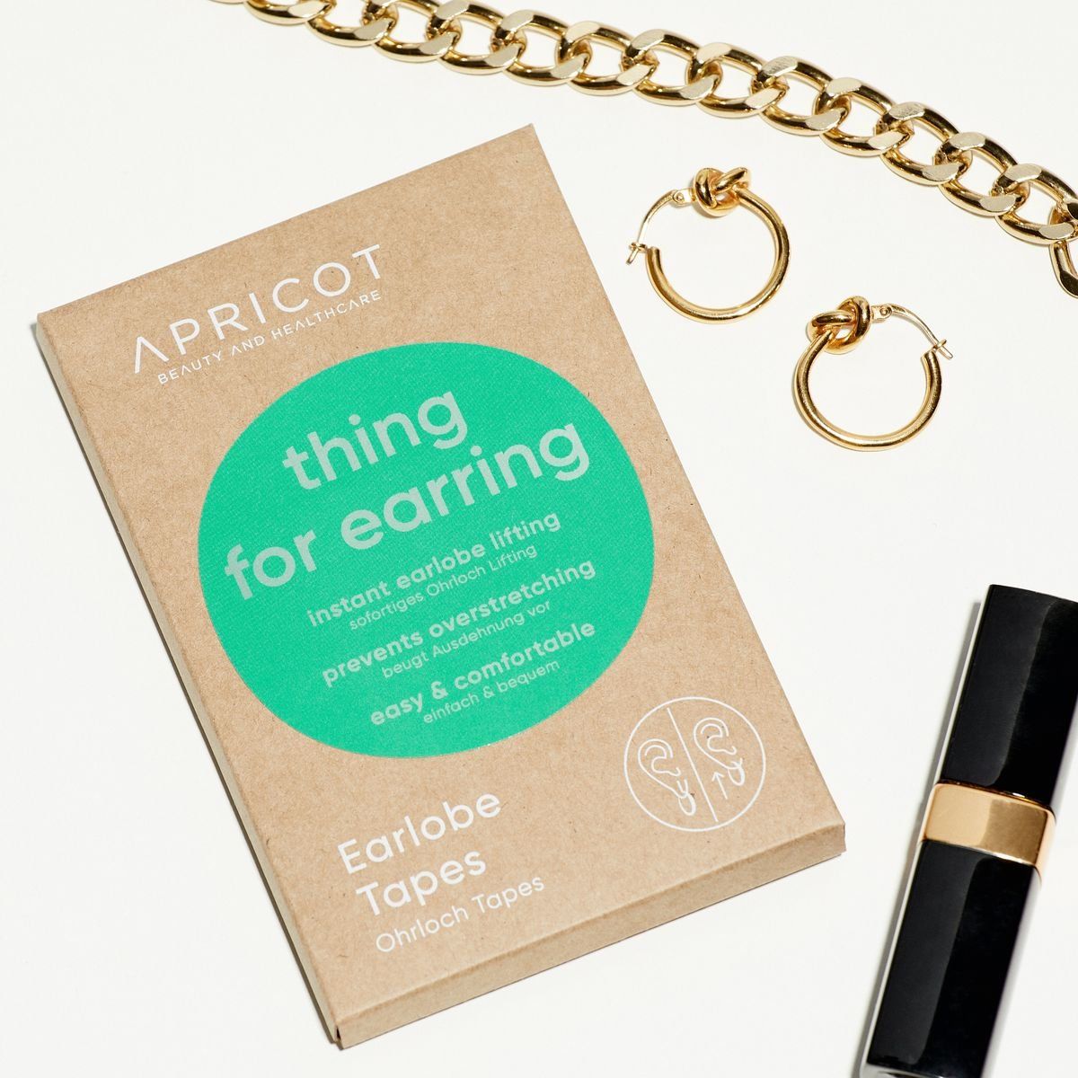 Confezione in cartone con testo del prodotto. Scritta: APRICOT, thing for earring, Ohrloch Tapes. Gioielli e cosmetici accanto.