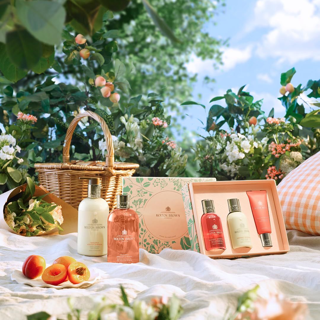 Scena picnic con prodotti Molton Brown. Cesto, coperta, frutta, confezione regalo e flaconi. Ambiente naturale.