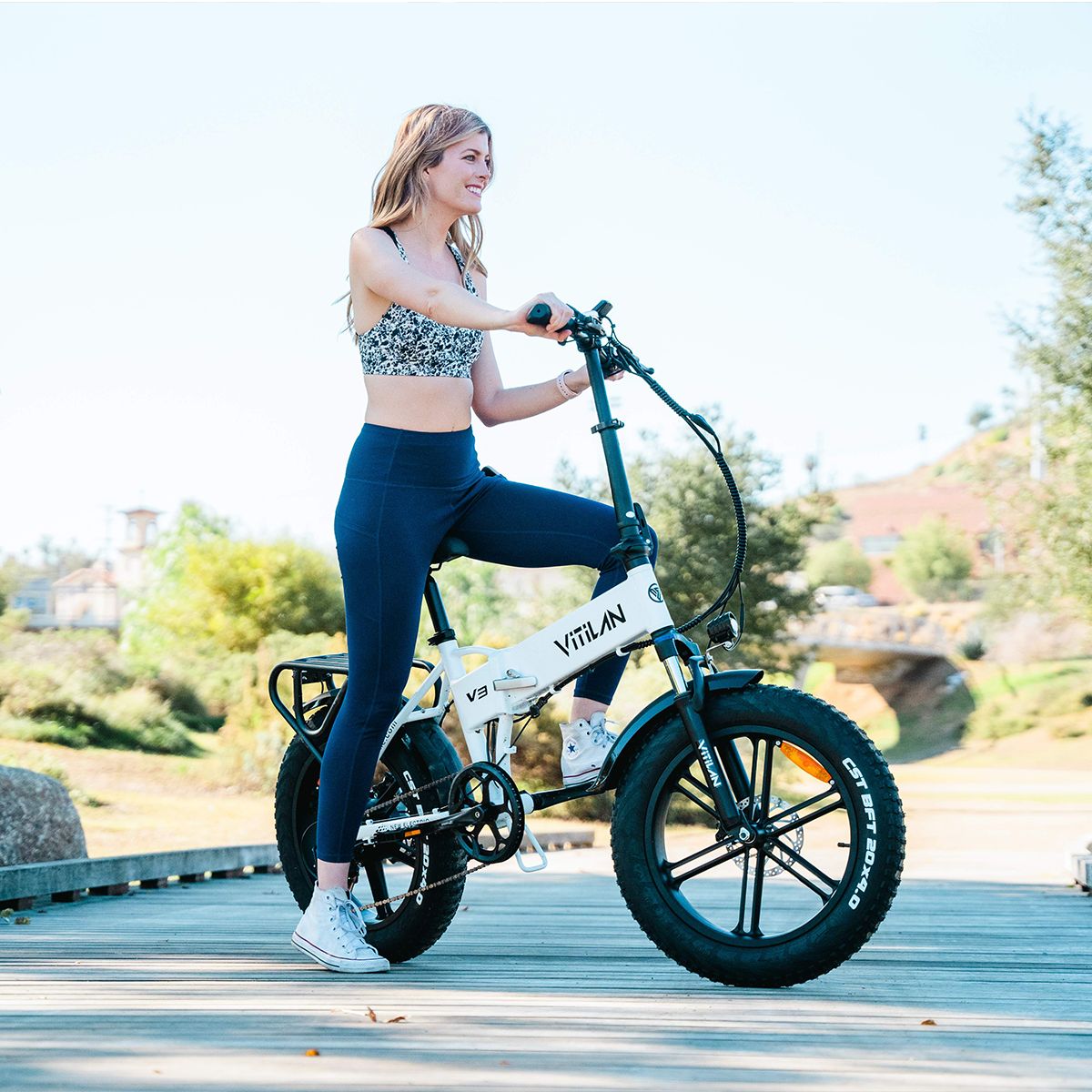 E-bike pieghevole bianco con pneumatici neri. Una donna in bicicletta su un ponte. Sul telaio è scritto Vitilan V3.