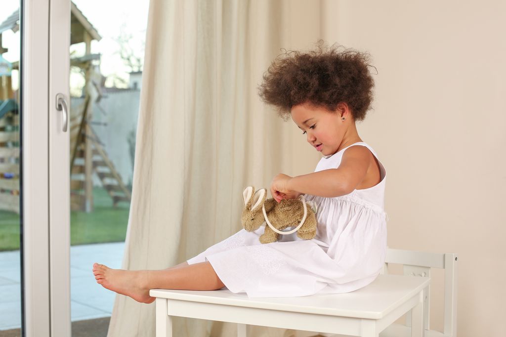 Una bambina in abito bianco seduta su un tavolo tiene in mano un coniglio di stoffa con corda.