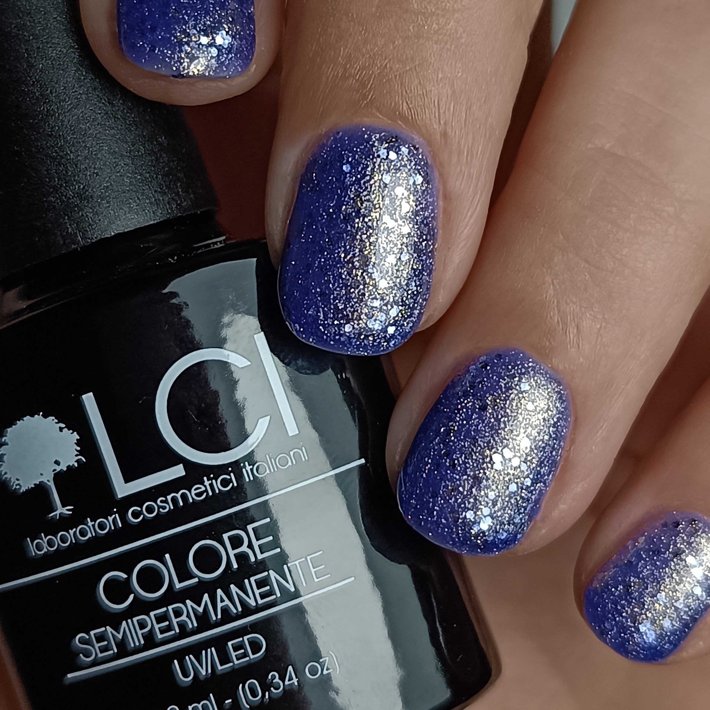 Unghie smaltate con smalto glitterato blu scuro. Flacone di smalto sullo sfondo. Scritta: LCI, Colore Semipermanente, UV/LED.