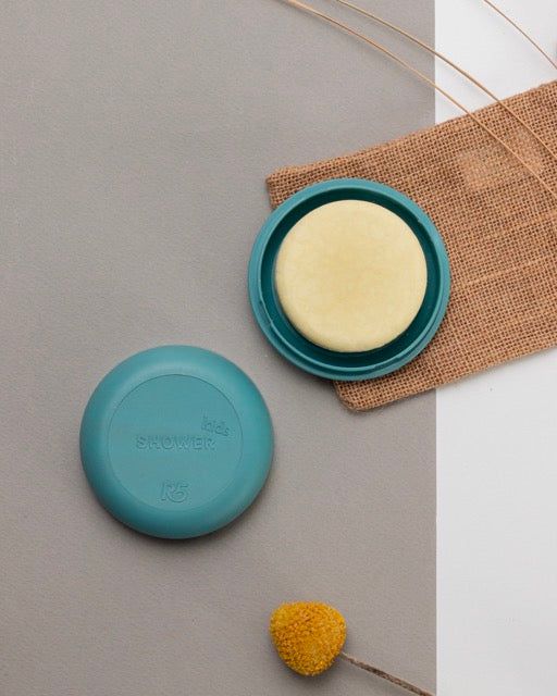 Contenitore rotondo turchese con docciaschiuma solido giallo. Coperchio con scritta « Shower ». Su tessuto.