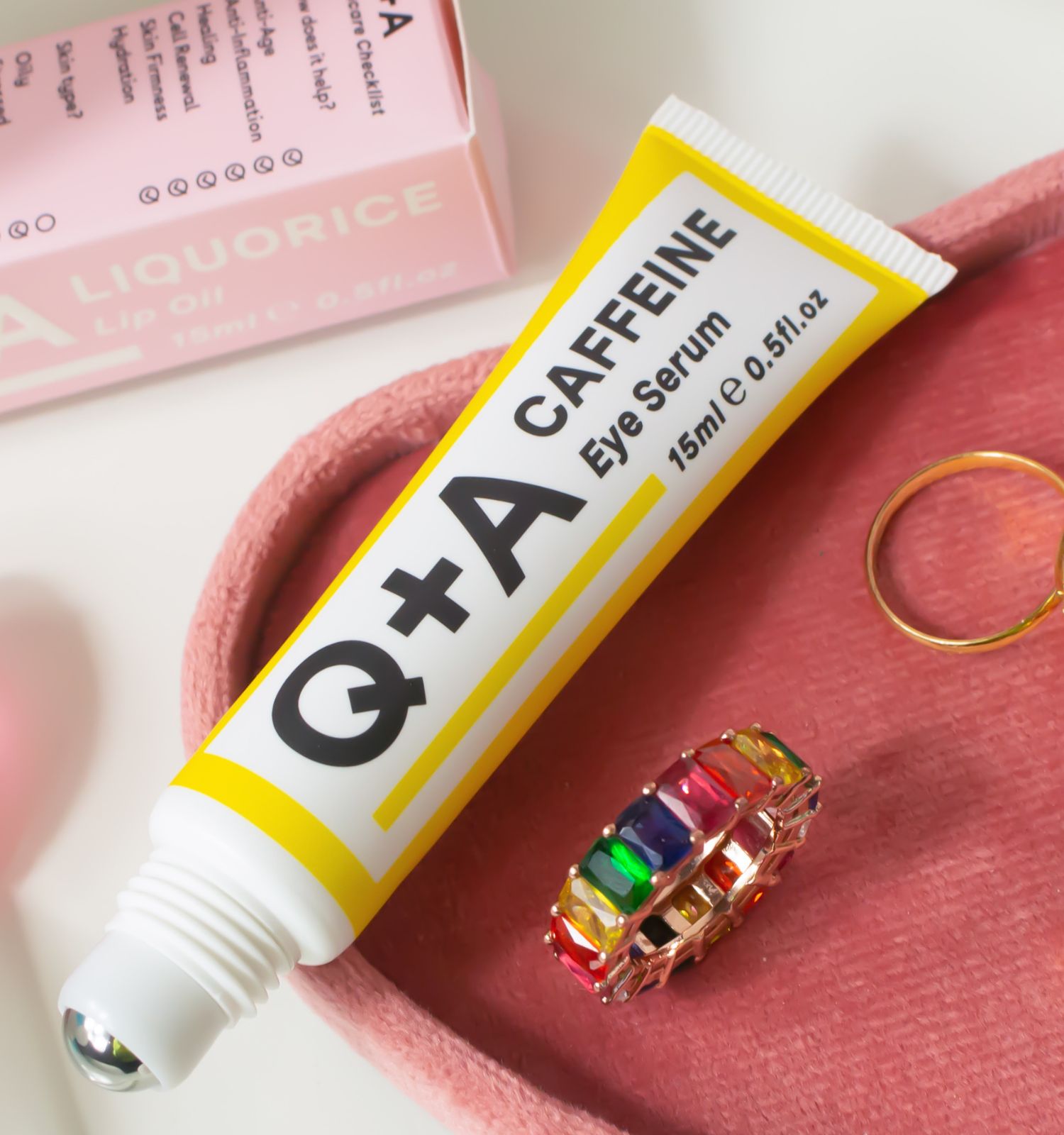 Tubo giallo con testo. Scritta: Q+A, CAFFEINE Eye Serum, 15ml 0.5fl.oz. Su sfondo rosa con gioielli.