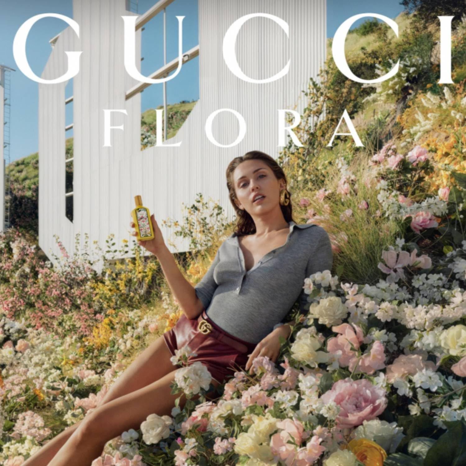 Donna tiene in mano un flacone di profumo giallo con design floreale. Scritta GUCCI FLORA sullo sfondo.