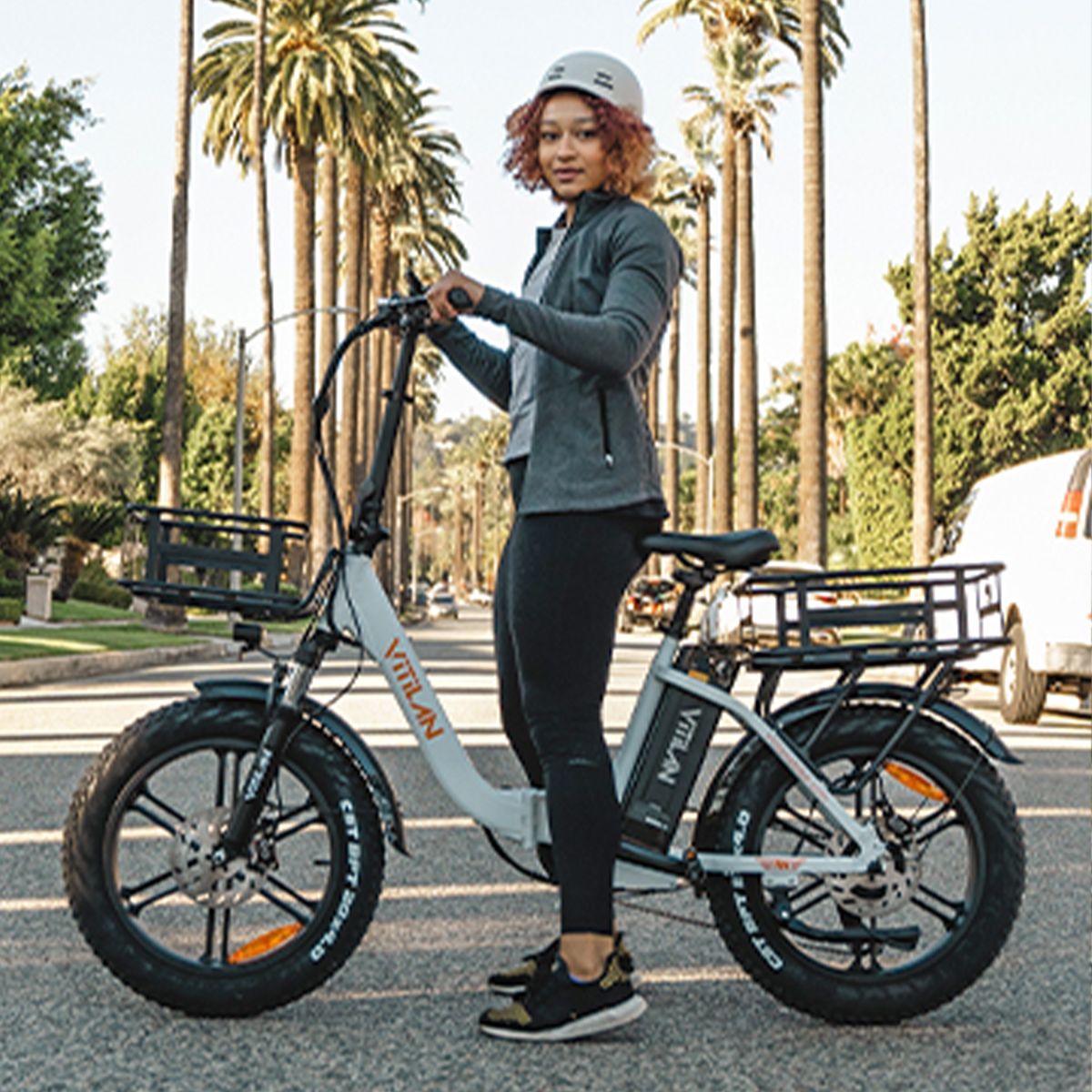Persona in piedi accanto a una bici elettrica Vitilan bianca. Indossa casco e abbigliamento sportivo. Palme sullo sfondo.