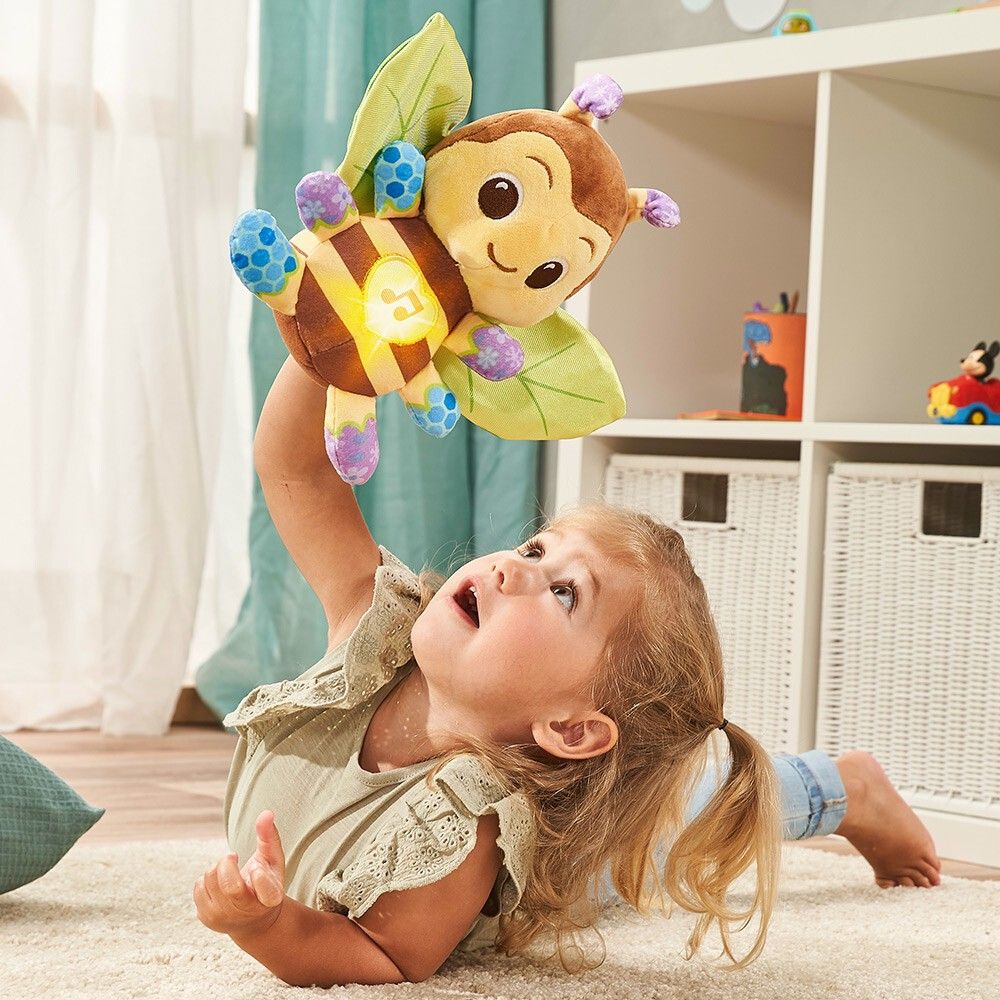 Bambina tiene in alto Vtech Musik-Kuschelbiene. L'ape è marrone, gialla e verde. La bambina è a terra.