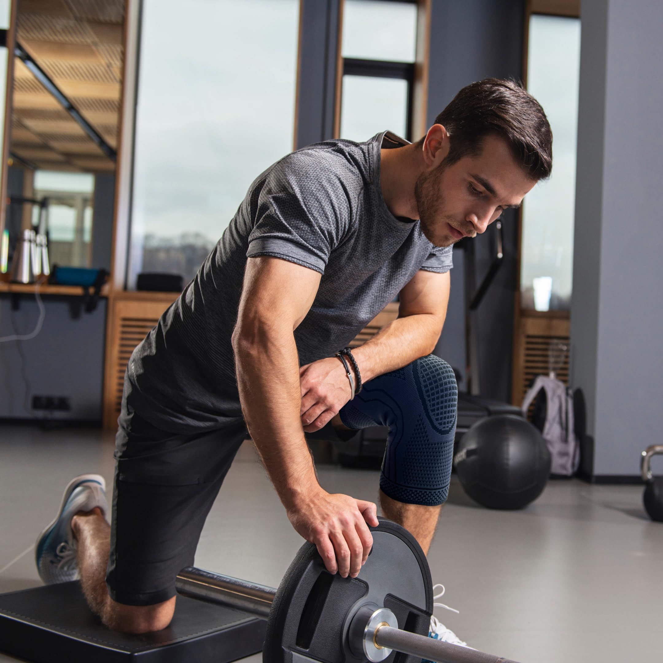 Uomo con ginocchiera durante l'allenamento. Si inginocchia e solleva un disco. Ambiente palestra.