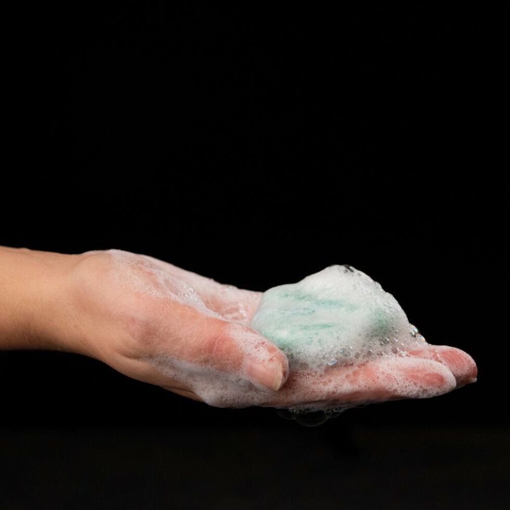 Mano con schiuma di sapone e barra shampoo verde. Sfondo nero.