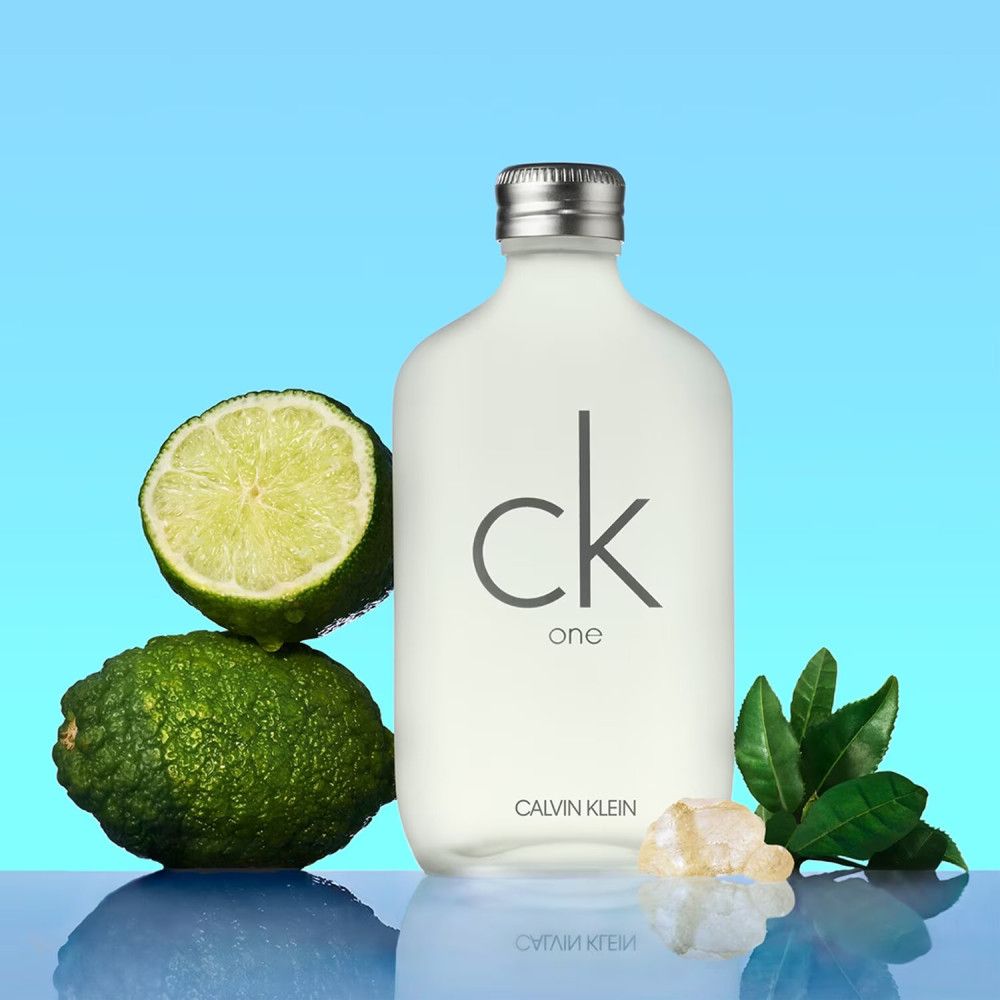 Flacone di profumo bianco con tappo argentato. Lime verdi e foglie. Scritta: ck one, Calvin Klein.