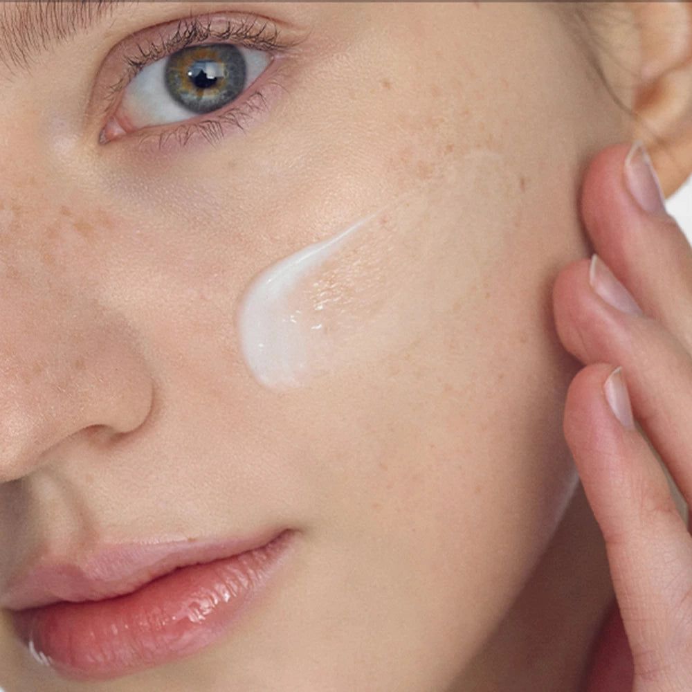 Primo piano di un viso. Una persona applica la crema sulla guancia. La pelle è chiara e ha lentiggini.