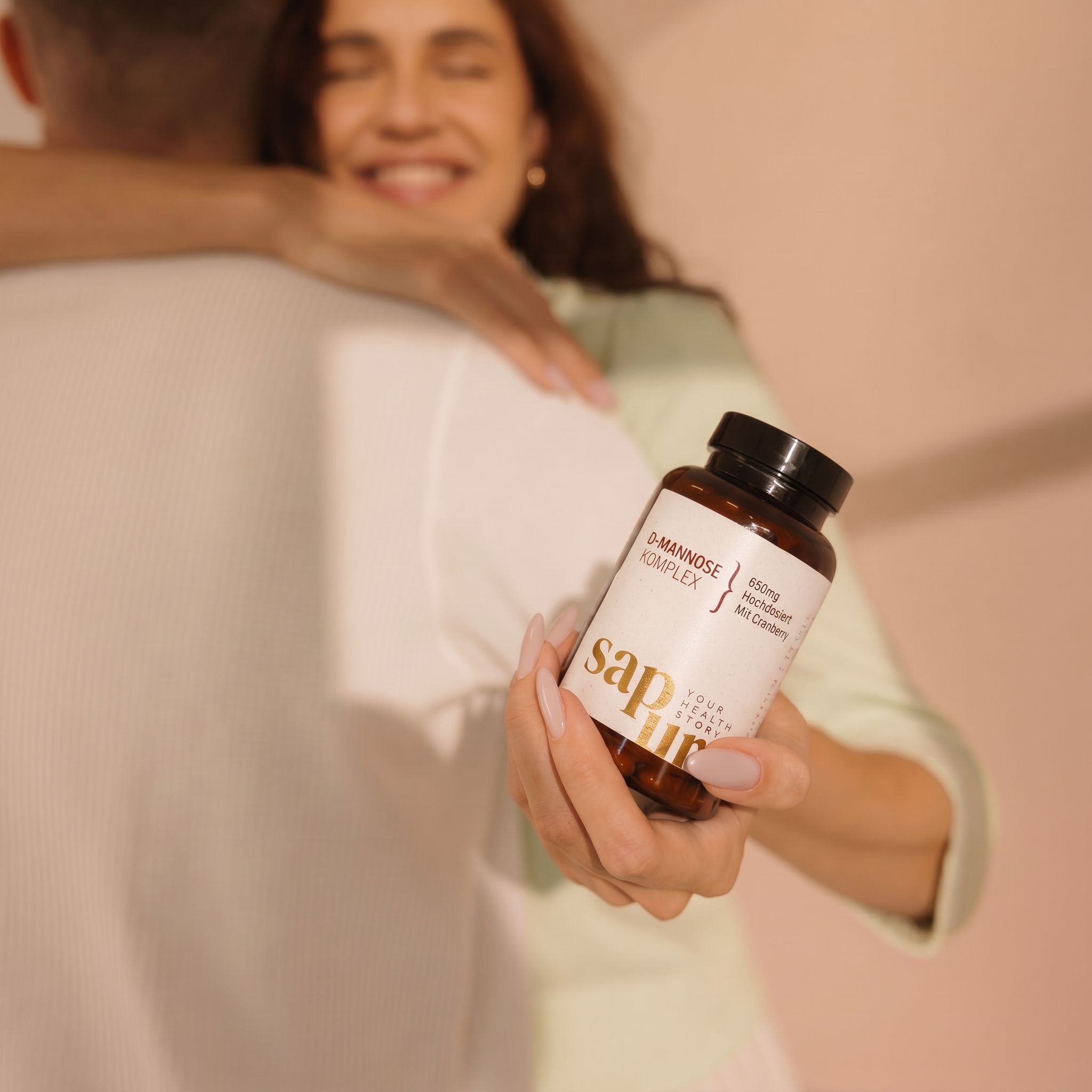 Una donna abbraccia un uomo, tenendo una bottiglia di D-Mannose Komplex+ 650mg con Cranberry.
