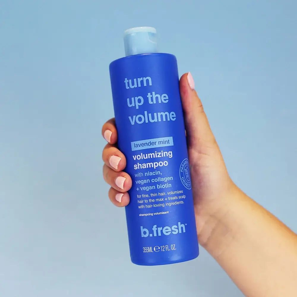 Flacone di shampoo blu "Turn Up The Volume" di b.fresh. Contiene niacina, collagene vegano e biotina. Tenuto in mano.