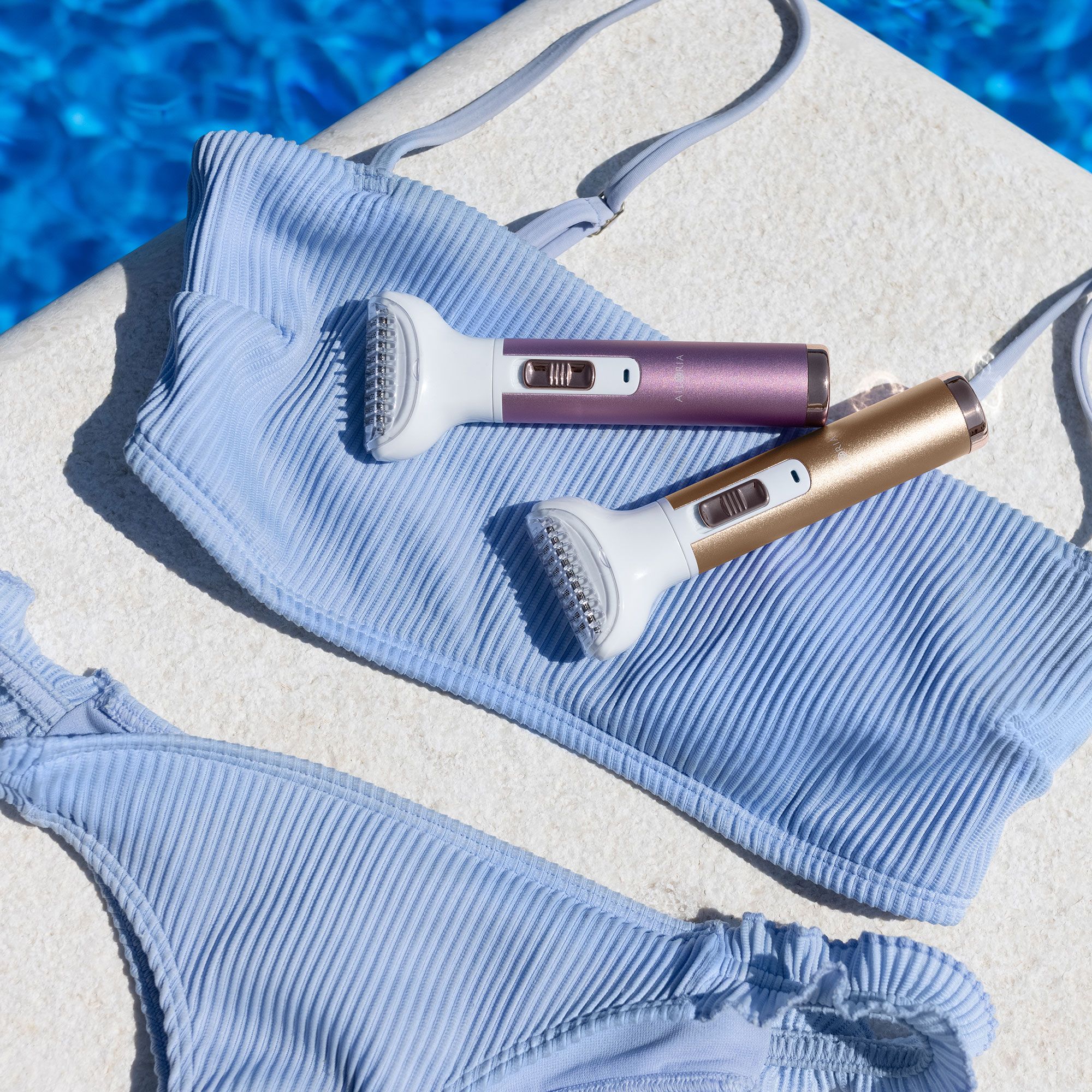 Due rasoi, rosa e dorato, appoggiati su un bikini azzurro. Acqua blu sullo sfondo.