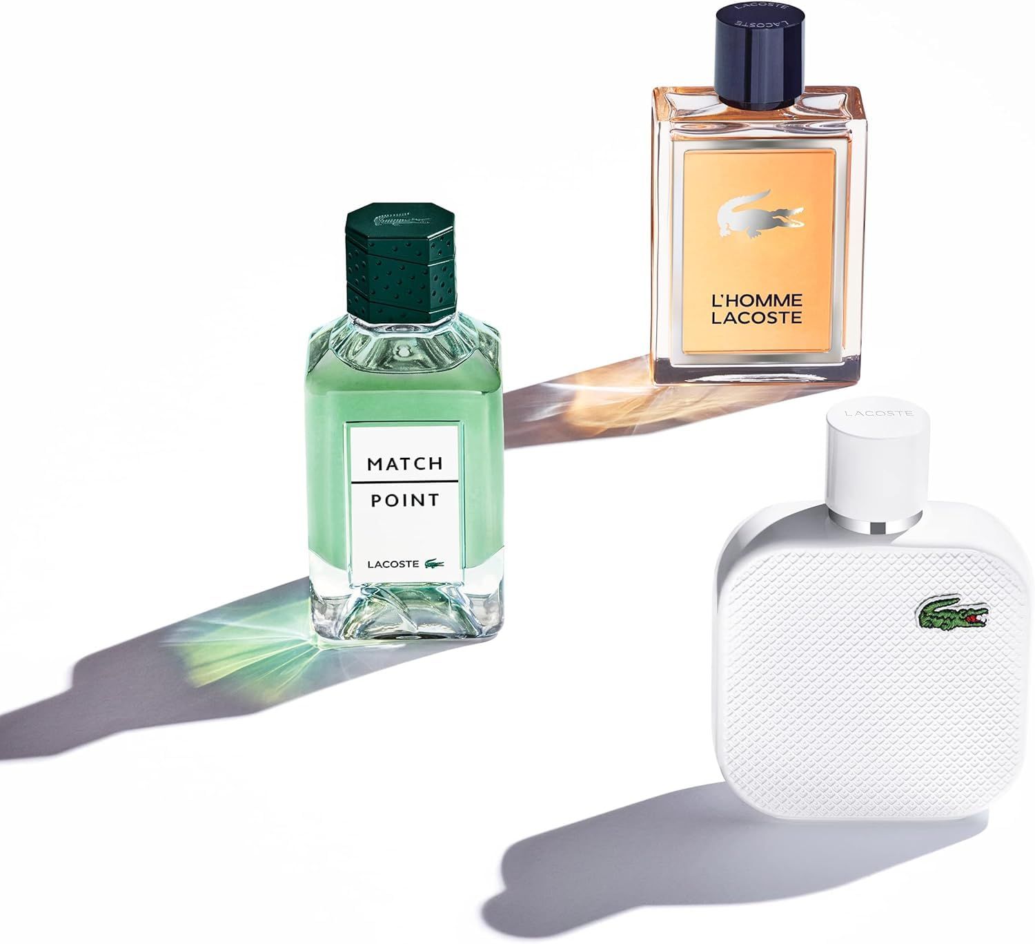 Tre flaconi di profumo. L'Homme Lacoste, Match Point e un flacone bianco con logo coccodrillo. Su sfondo bianco.