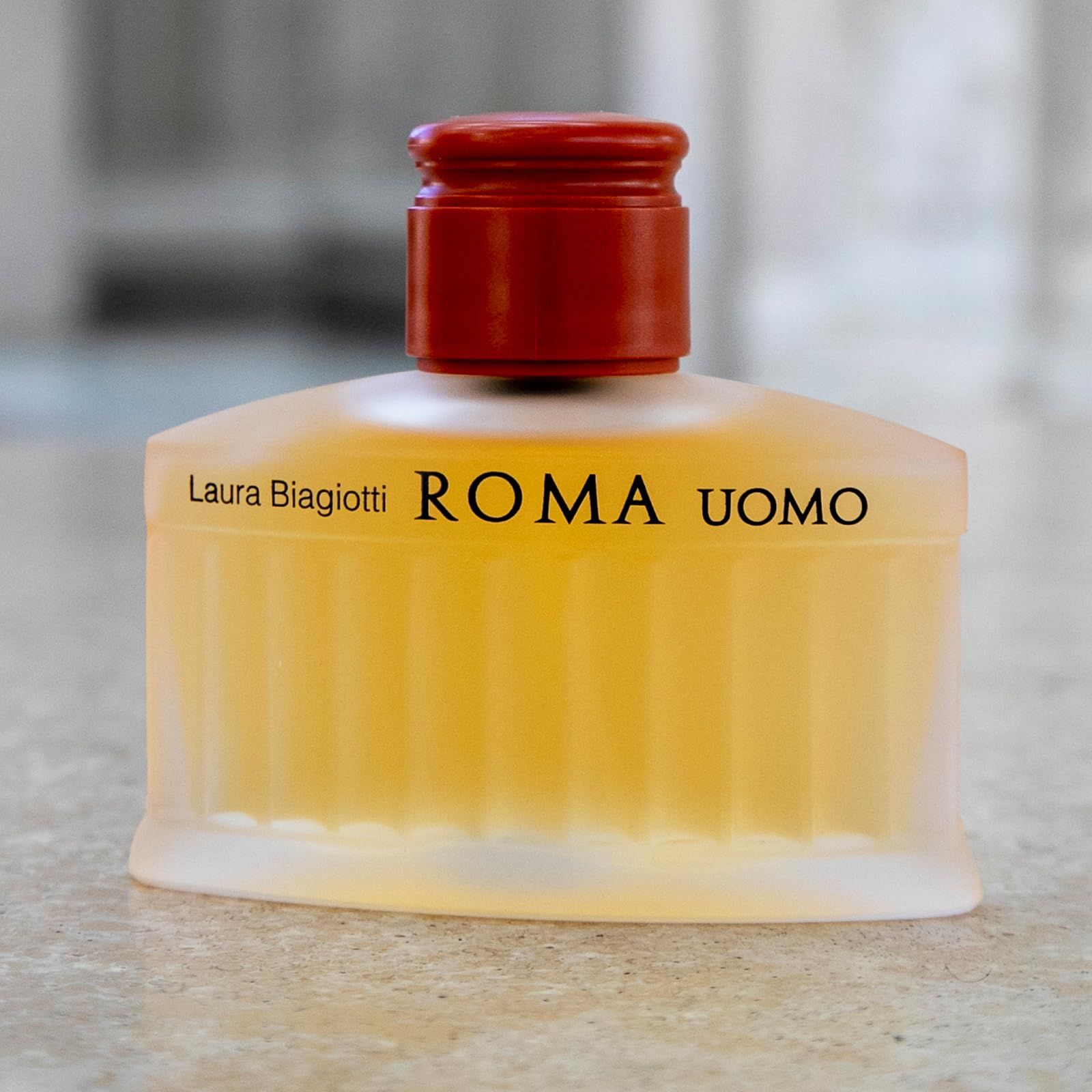 Flacone di profumo con liquido giallastro. Scritta: Laura Biagiotti ROMA UOMO. Su sfondo grigio.