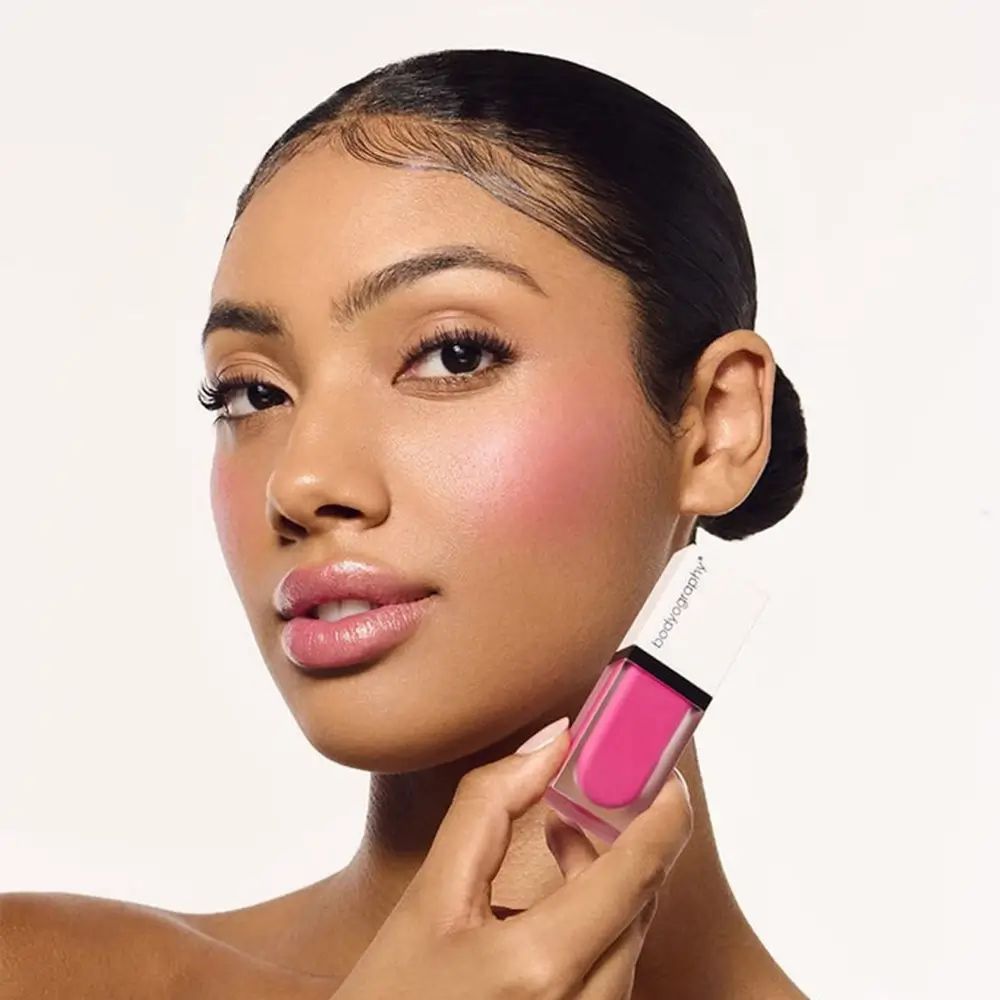 Donna con fard. Flacone di fard liquido BODYOGRAPHY in mano. Colore fucsia.