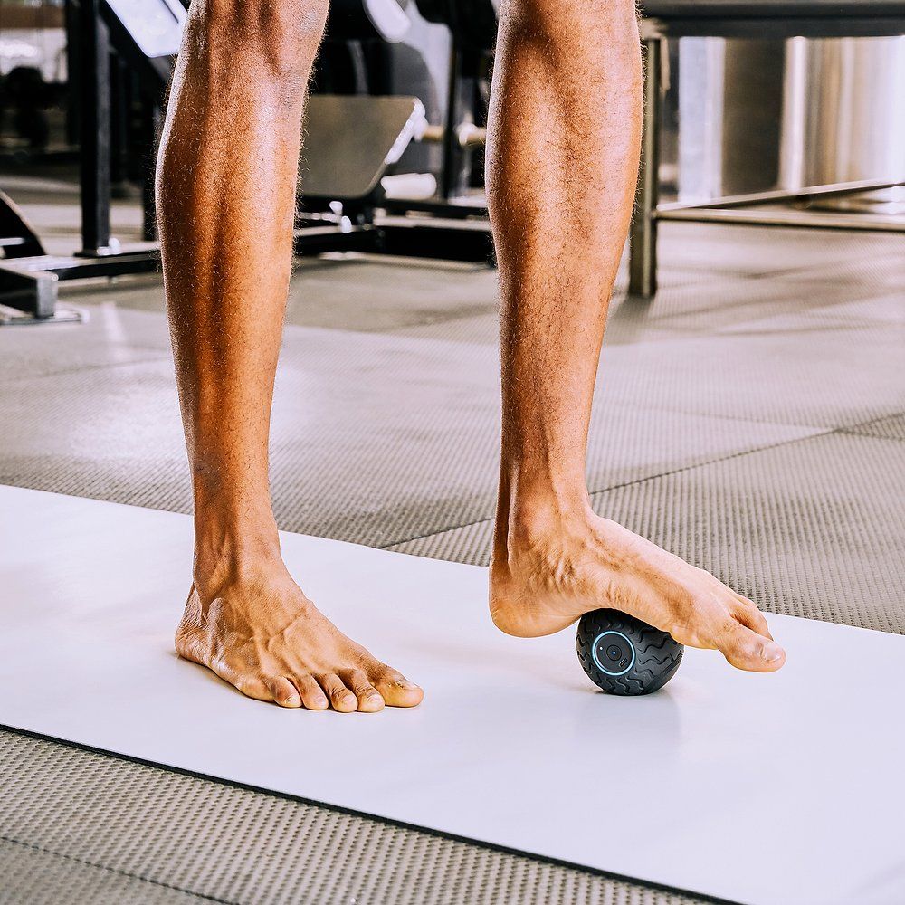 Piedi su un tappetino, una sfera da massaggio nera sotto il piede. Ambiente palestra.