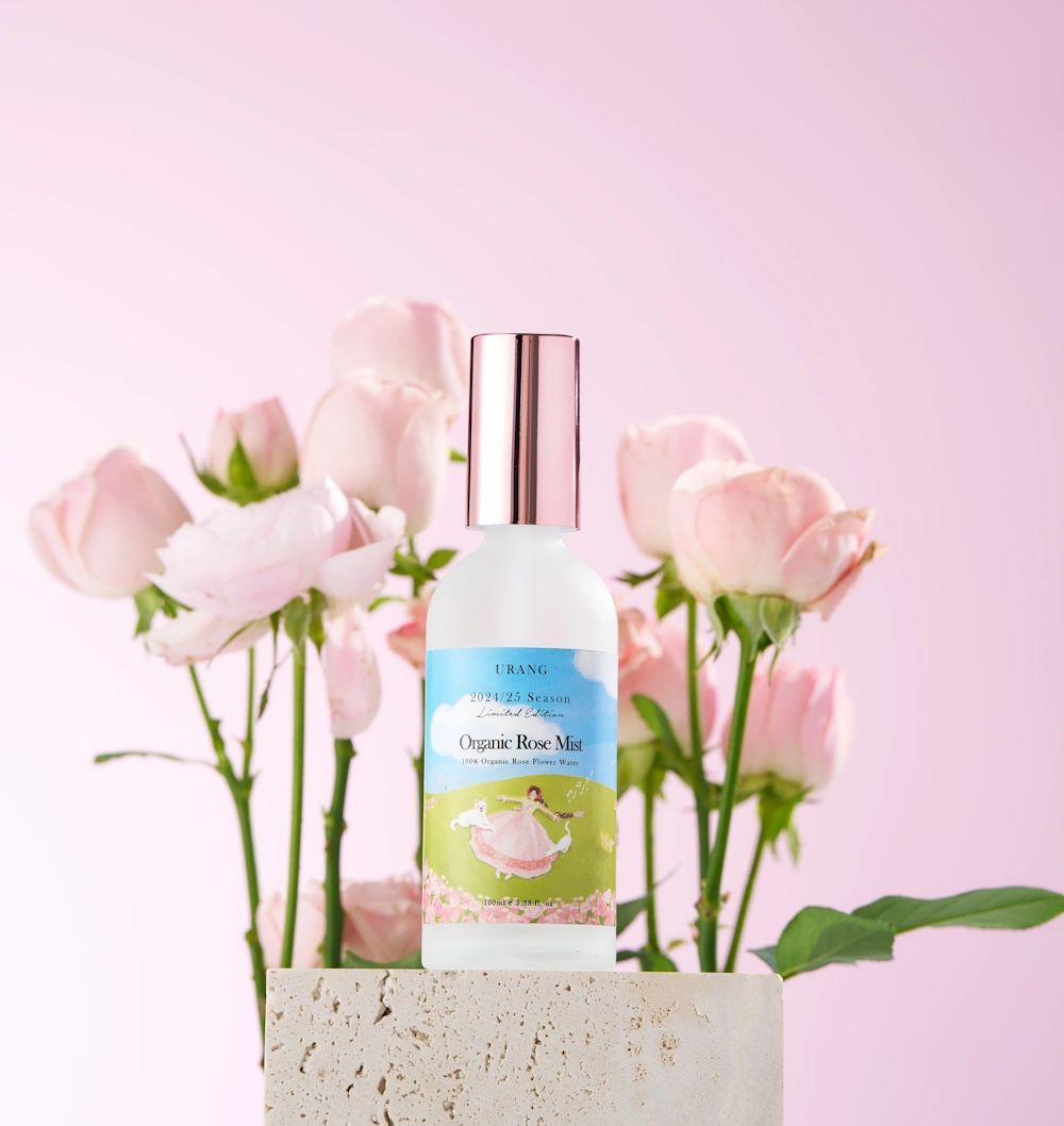 Flacone spray con tappo color oro rosa. Etichetta con testo e illustrazione. Davanti a rose rosa.