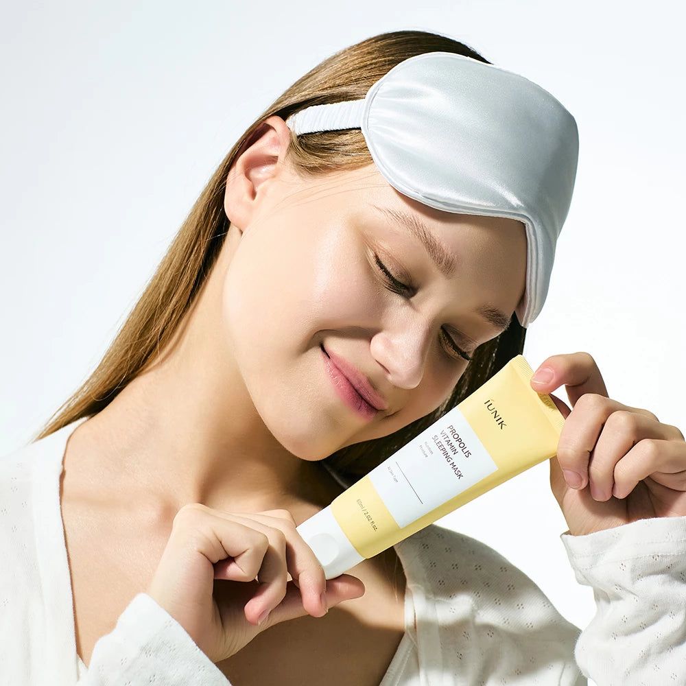Donna con mascherina per dormire tiene un tubo giallo. Scritta: IUNIK, PROPOLIS VITAMIN SLEEPING MASK. Sorride.